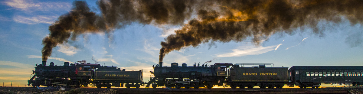 Two steam locomotives silhouetted against a sunset as they cross a low brige